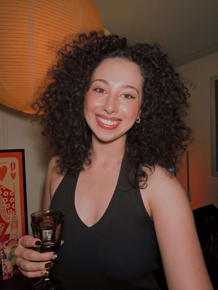 A woman with curly brown hair smiling at the camera while holding a glass in her hand.